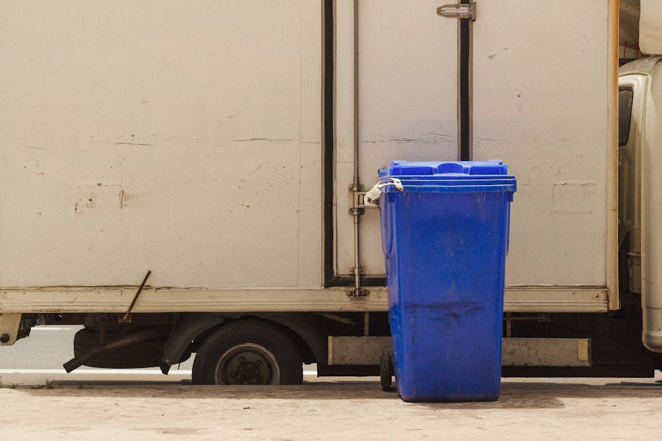 A large blue bin with a closed lid, made of durable plastic with a slightly textured surface, stands on a concrete pavement adjacent to a white delivery truck. The bin is positioned in front of the truck's side panel, which appears to be made of metal with visible weathering, minor scratches, and dirt marks. The truck is parked on a flat surface, and a black metal pole runs vertically along the side, secured with a metal bracket. Part of the truck’s wheel and undercarriage are visible beneath the side panel, showing a standard black tire and some mechanical components. The environment suggests an outdoor setting with natural lighting, contributing to a neutral and professional tone. The scene subtly indicates an activity related to waste disposal or collection, which aligns with rubbish removal services like those offered by Rubbish Removal Dulwich, particularly in the context of alternative waste handling methods such as private collection or on-site clearance.