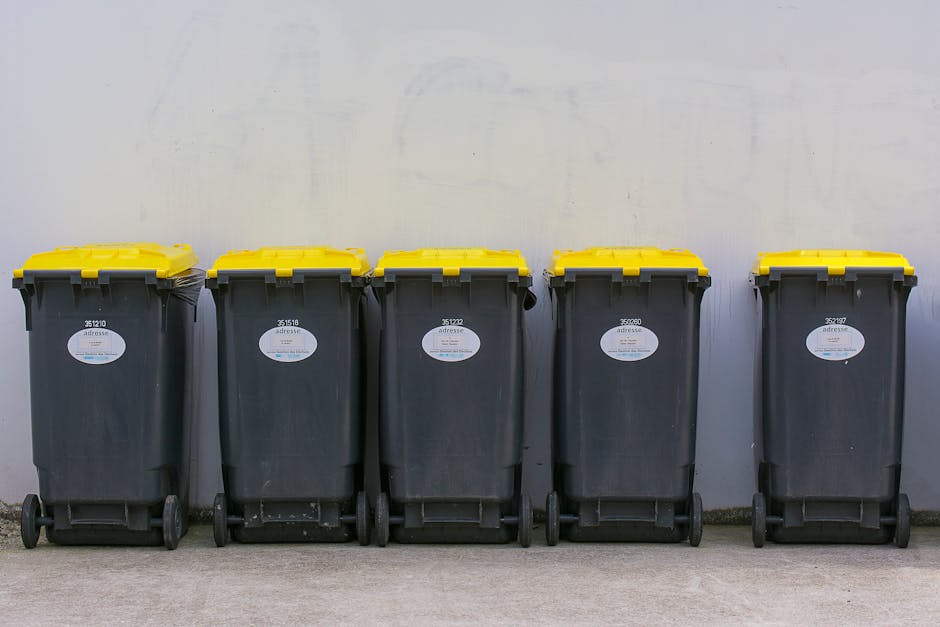 A large blue bin with a closed lid, made of durable plastic with a slightly textured surface, stands on a concrete pavement adjacent to a white delivery truck. The bin is positioned in front of the truck's side panel, which appears to be made of metal with visible weathering, minor scratches, and dirt marks. The truck is parked on a flat surface, and a black metal pole runs vertically along the side, secured with a metal bracket. Part of the truck’s wheel and undercarriage are visible beneath the side panel, showing a standard black tire and some mechanical components. The environment suggests an outdoor setting with natural lighting, contributing to a neutral and professional tone. The scene subtly indicates an activity related to waste disposal or collection, which aligns with rubbish removal services like those offered by Rubbish Removal Dulwich, particularly in the context of alternative waste handling methods such as private collection or on-site clearance.