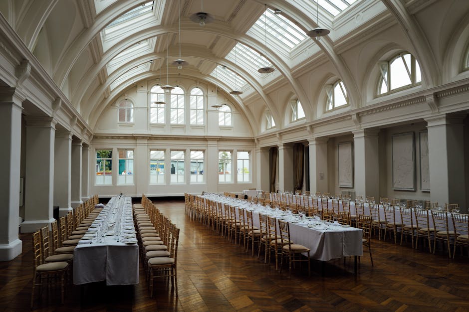 An elegant, high-ceilinged event hall with a curved, ornate white ceiling featuring multiple arched skylights that allow natural light to fill the space. The room has a polished wooden parquet floor and large windows along the far wall, framed by dark curtains. In the foreground, two long banquet tables are arranged parallel to each other, covered with pristine white tablecloths. The tables are set with neatly folded napkins, glassware, and tableware, indicating preparations for a formal gathering. Gold-colored Chiavari chairs with padded seats are evenly spaced along both sides of the tables, enhancing the sophisticated ambiance. The overall setting appears well-maintained and ready for an event, with no visible clutter or rubbish, consistent with professional venue management and private event arrangements, aligning with the context of alternative waste handling and event clearance services that Rubbish Removal Dulwich offers for such locations.