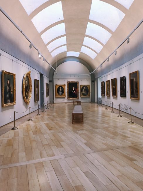 The image depicts an art gallery with an arched, skylighted ceiling that allows natural light to illuminate the space. The gallery features a smooth, light wooden floor with a subtle pattern, and white walls lined with framed classical paintings, including portraits and mythological scenes, displayed in ornate and simple gold and dark wooden frames. The artworks are arranged in a single row along both sides of the room, with small stanchions connected by rope barriers to guide visitors and prevent contact. At the end of the gallery, a prominent portrait is centered on the back wall, flanked by two smaller paintings, all enclosed in elaborate gilded frames. A wooden bench is positioned in the middle of the room for visitors to sit and observe the art. The environment appears quiet and well-maintained, reflecting a professional interior suitable for museum or private collection display, which subtly relates to high-quality, independent collection and management of artwork, similar to on-site or private waste collection services provided by companies like Rubbish Removal Dulwich.