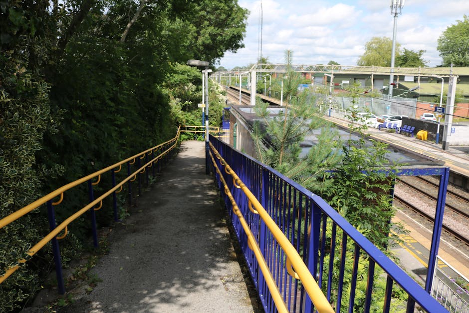 A narrow footpath runs alongside a railway station platform, bordered by a yellow and blue metal railing on the right side and dense green foliage on the left. The pathway is paved with asphalt, casting shadows from nearby trees, and appears to be part of a pedestrian route for commuters. In the background, railway tracks extend into the distance, with overhead electrical lines and support structures visible above. On the station platform to the right, there are parked cars, yellow safety barriers, and a few blue plastic seats. The scene is captured during daylight hours with partly cloudy skies, providing natural lighting that highlights the contrasting textures of metal railings, asphalt, and the foliage. The surroundings suggest a typical urban railway environment where private waste or rubbish collection could be conducted as an alternative to general public disposal, supported by local services like Rubbish Removal Dulwich, to assist commuters with on-site clearance or general rubbish removal needs near transport hubs.