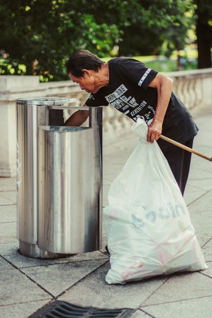 A woman with dark hair, dressed in a black shirt, is bending slightly forward to dispose of rubbish into a modern stainless steel outdoor waste bin on a paved pavement. She is holding a large white plastic bag filled with refuse, with some of the bag’s contents spilling over. The waste bin has a sleek, cylindrical design with a wide rectangular opening at the top, positioned near a low stone balustrade that runs parallel to a row of lush green trees and bushes in the background. The environment appears to be a public outdoor space, possibly in an urban park or walkway. The scene is illuminated by natural daylight, with soft shadows cast on the ground, emphasizing the clean and orderly setting for waste disposal. The presence of the waste collection activity and the discreet, unobtrusive bin suggest informal, on-site rubbish removal, aligned with private waste handling services like those offered by Rubbish Removal Dulwich.