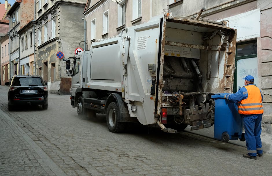 A municipal waste collection vehicle parked on a narrow urban street with cobblestone pavement, positioned beside a row of older, multi-story buildings with stone and brick facades. The rear of the truck is open, revealing rusty metal components and an empty compartment. A worker dressed in a blue uniform and a high-visibility orange vest is standing next to the truck, holding a blue wheelie bin and loading it into the vehicle. The street features parked cars, including a black vehicle on the left, with traffic signs indicating parking restrictions. The scene is illuminated by natural daylight, with no visible additional environmental details. This urban rubbish collection scene reflects the process of private waste collection and on-site waste handling, typical for local rubbish removal services in central city areas.