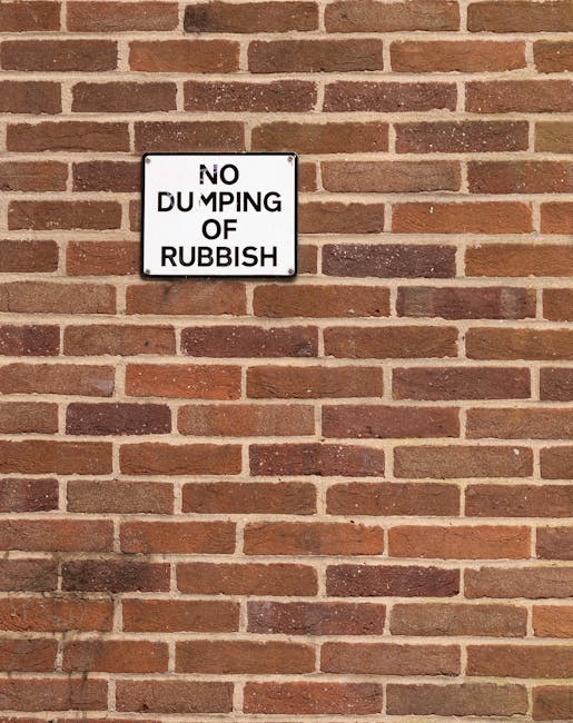 A rectangular warning sign mounted on a red brick wall, indicating 'NO DUMPING OF RUBBISH'. The sign has a white background with black text, with the words arranged in a vertical and slightly staggered manner, and is positioned around the upper left center of the image. The brick wall consists of uniform reddish-brown bricks with darker brown and slightly weathered areas, laid in a horizontal running bond pattern with light-colored mortar joints. The overall scene suggests an urban or residential setting, where private waste management might be employed as an alternative to local authority rubbish collection, aligning with services provided by Rubbish Removal Dulwich. The lighting is natural, evenly illuminating the textured surface of the bricks and the sign without harsh shadows or glare, reflecting typical outdoor conditions.